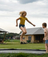 Dos niños juegan en una cama elástica frente a tiendas glamping en un parque vacacional rural.