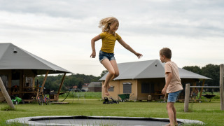 Dos niños juegan en una cama elástica frente a tiendas glamping en un parque vacacional rural.