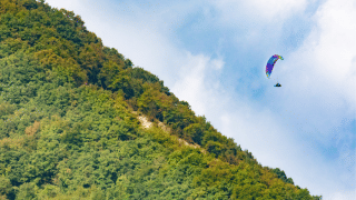 Un parapente colorido vuela sobre una colina boscosa bajo un cielo azul en Veneto, Italia.