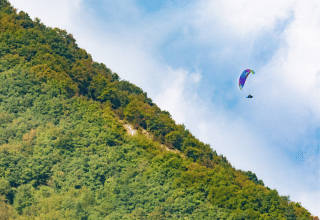 Un parapente colorido vuela sobre una colina boscosa bajo un cielo azul en Veneto, Italia.