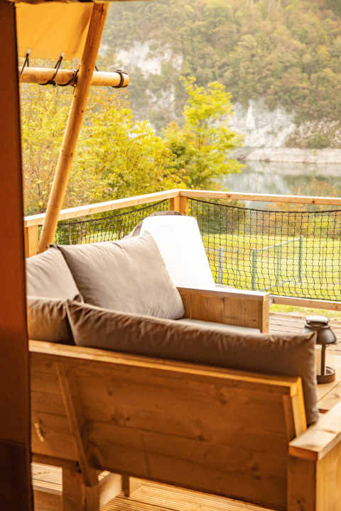 Divano di legno con cuscini su terrazza di tenda safari al Camping Gajole in Italia, vista lago e bosco.