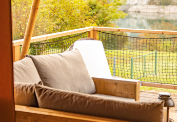 Wooden sofa with cushions on a safari tent deck at Camping Gajole in Italy, overlooking lake and forest.