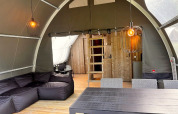 Intérieur de la tente safari Big Oak Lodge avec canapé noir, table à manger, murs en bois et luminaires suspendus.