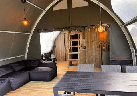 Interno della tenda safari Big Oak Lodge con divano nero, tavolo da pranzo, parete in legno e lampade a sospensione.