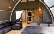 Interior de la tienda safari Big Oak Lodge con sofá negro, mesa de comedor, pared de madera y lámparas colgantes.