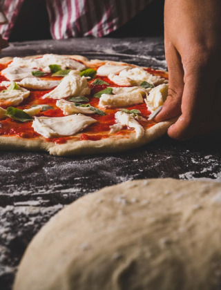 Persona preparando una pizza casera con salsa de tomate, queso y albahaca sobre una superficie enharinada.