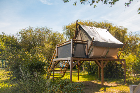 Tienda safari elevada en una plataforma de madera rodeada de naturaleza verde, tomada de día.