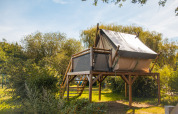 Tienda safari elevada en una plataforma de madera rodeada de naturaleza verde, tomada de día.