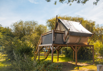 Tente safari surélevée sur une plateforme en bois, entourée de végétation verdoyante sous le ciel bleu.