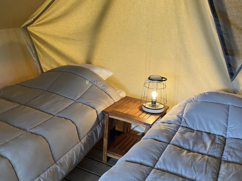 Dos camas con ropa de cama gris en una tienda tipi, separadas por una mesa con lámpara, ambiente acogedor.