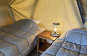 Two beds with gray bedding inside a teepee tent, separated by a small table holding a lamp, cozy setup.