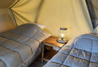 Dos camas con ropa de cama gris en una tienda tipi, separadas por una mesa con lámpara, ambiente acogedor.