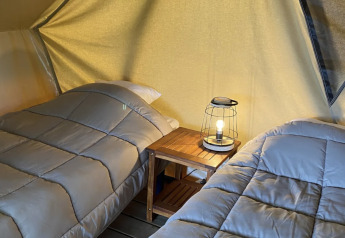 Two beds with gray bedding inside a teepee tent, separated by a small table holding a lamp, cozy setup.