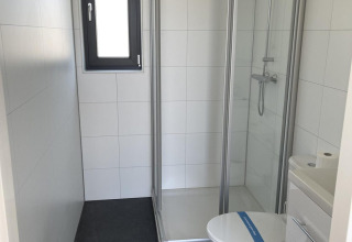 Baño moderno de glamping con cabina de ducha, inodoro y lavabo pequeño, paredes blancas y suelo oscuro.