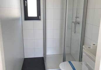 Baño moderno de glamping con cabina de ducha, inodoro y lavabo pequeño, paredes blancas y suelo oscuro.