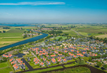 Luchtbeeld van een glampingverblijf met rode dakjes, grachten en groene velden onder een blauwe lucht.