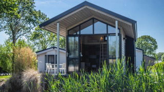 Moderne Tiny House-lodge met grote ramen, terrasstoelen en omringd door groene, natuurlijke begroeiing.