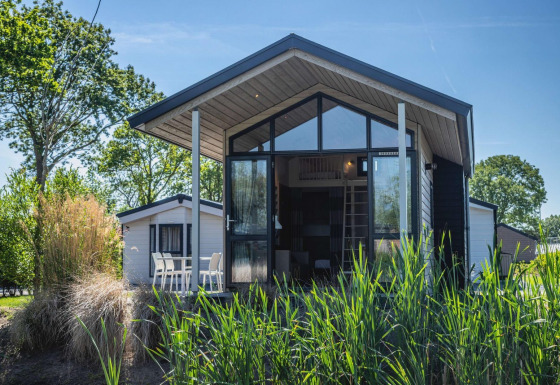 Moderna Tiny House lodge con grandi finestre in vetro, area salotto esterna e vegetazione rigogliosa intorno.