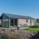 Moderne træhytte ved vandkant med terrasse og havemøbler ved Velthorst Lodge under klar blå himmel.