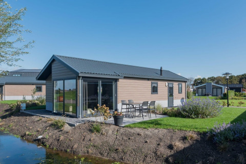 Modernes Holzlodge Velthorst mit Terrasse, Gartenmöbeln und Glasfront an einem kleinen Wasserlauf.