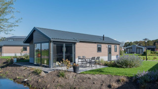 Modernes Holzlodge Velthorst mit Terrasse, Gartenmöbeln und Glasfront an einem kleinen Wasserlauf.