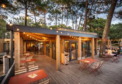 Terraza al aire libre con guirnaldas de luces y mesas de madera en Huttopia Arcachon, Nouvelle-Aquitania, Francia.