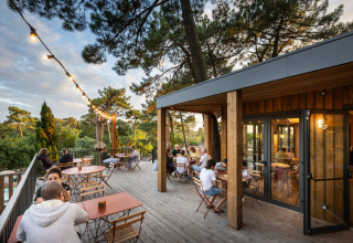 Terrasse extérieure du parc de vacances Huttopia Arcachon avec clients, arbres et guirlandes lumineuses.