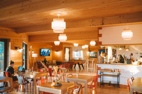 Vista interna di una caffetteria accogliente a Huttopia Arcachon, Francia, con arredi in legno e atmosfera calda.