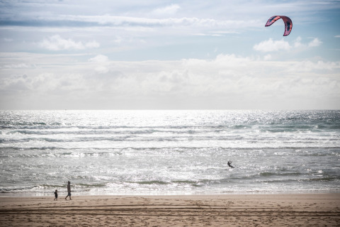 Escena de playa en Huttopia Arcachon, Nouvelle-Aquitaine, Francia, con personas y un kitesurfista en el mar.