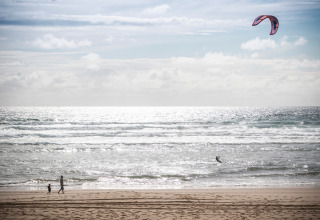 Scène de plage à Huttopia Arcachon en Nouvelle-Aquitaine, France, avec promeneurs et kitesurfeur en mer.