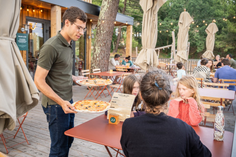 Un camarero sirve pizza a una familia en la terraza del parque vacacional Huttopia Arcachon en Francia.
