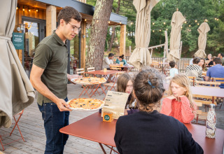Een ober serveert pizza aan een gezin op het terras van het Huttopia Arcachon vakantiepark in Frankrijk.