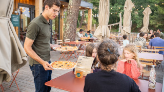 Un camarero sirve pizza a una familia en la terraza del parque vacacional Huttopia Arcachon en Francia.
