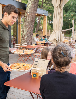 Un camarero sirve pizza a una familia en la terraza del parque vacacional Huttopia Arcachon en Francia.