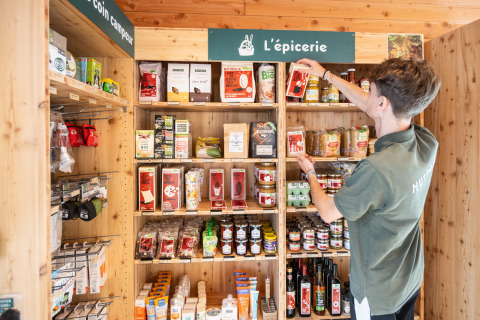 Persoon ordent boodschappen op houten rekken onder het bord 'L'épicerie' bij Huttopia Arcachon.