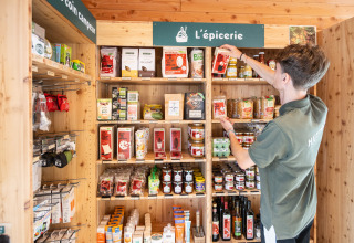 Persoon ordent boodschappen op houten rekken onder het bord 'L'épicerie' bij Huttopia Arcachon.