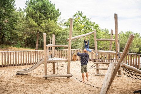 Aire de jeux en bois sur sable à Huttopia Arcachon, France, enfant grimpant, entouré d’arbres.