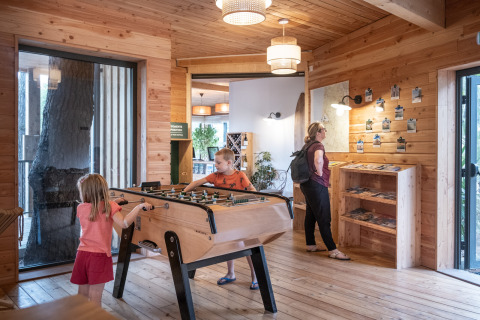 Interior de una cabaña de madera en Huttopia Arcachon, niños juegan futbolín, adulto revisa folletos.