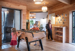 Interior of a wooden cabin at Huttopia Arcachon, kids playing foosball, adult looking at brochures on wall.