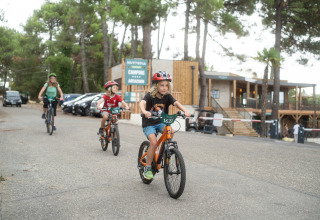 Des enfants et un adulte font du vélo devant le parc Huttopia Arcachon en Nouvelle-Aquitaine, France, entourés d’arbres.