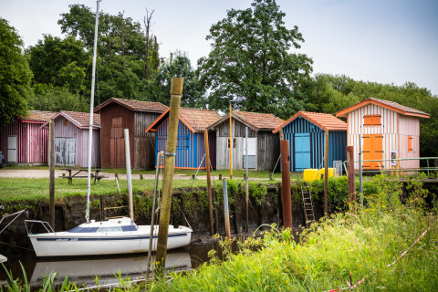 Bonte houten huisjes en een zeilboot langs de oever bij Arcachon, omgeven door weelderig groen.