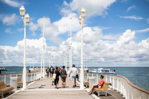 Personas pasean y descansan en bancos de un muelle soleado cerca de Arcachon, Francia, con vista al mar.