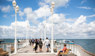 Personas pasean y descansan en bancos de un muelle soleado cerca de Arcachon, Francia, con vista al mar.