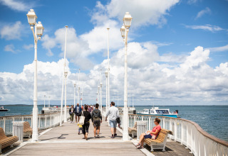 Personas pasean y descansan en bancos de un muelle soleado cerca de Arcachon, Francia, con vista al mar.