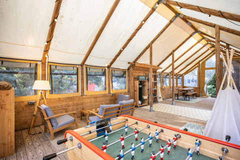 Interior acogedor de una tienda en Huttopia Oléron les Pins, con sofás, futbolín y ventanas grandes.