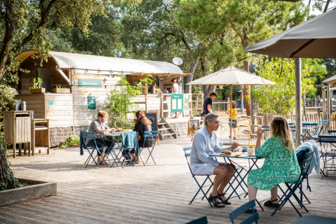 Gemütlicher Außenbereich eines Cafés im Huttopia Oléron les Pins, Menschen essen und entspannen im Grünen.
