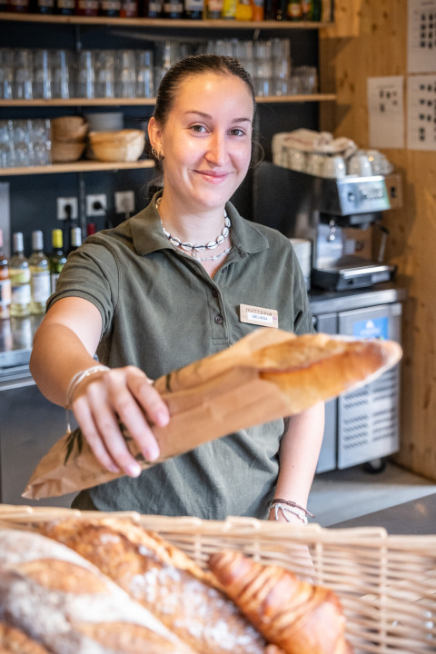 Empleada sonriente en Huttopia Oléron les Pins entrega una baguette en la panadería del parque vacacional.