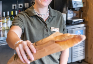 En smilende kvinde i polo, der arbejder i bageriet på Huttopia Oléron les Pins, rækker en baguette frem.