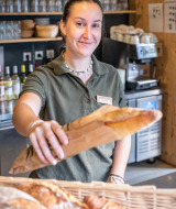 Empleada sonriente en Huttopia Oléron les Pins entrega una baguette en la panadería del parque vacacional.