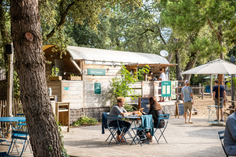 Zona de cafetería al aire libre en Huttopia Oléron les Pins, con personas comiendo entre árboles y naturaleza.